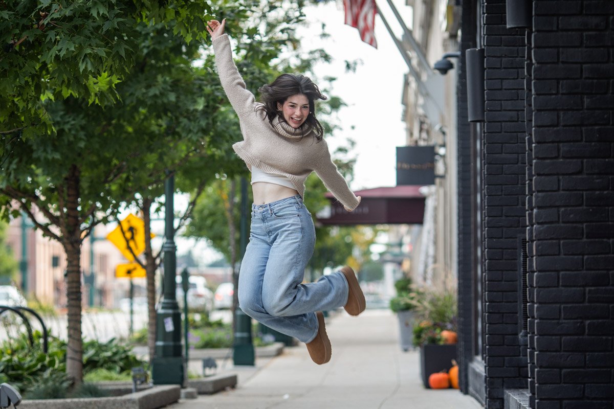 birmingham-senior-pictures-photography Senior portrait of a girl in Downtown Birmingham, Michigan, jumping high in the air in a street scene. Wearing blue jeans and a brown sweater