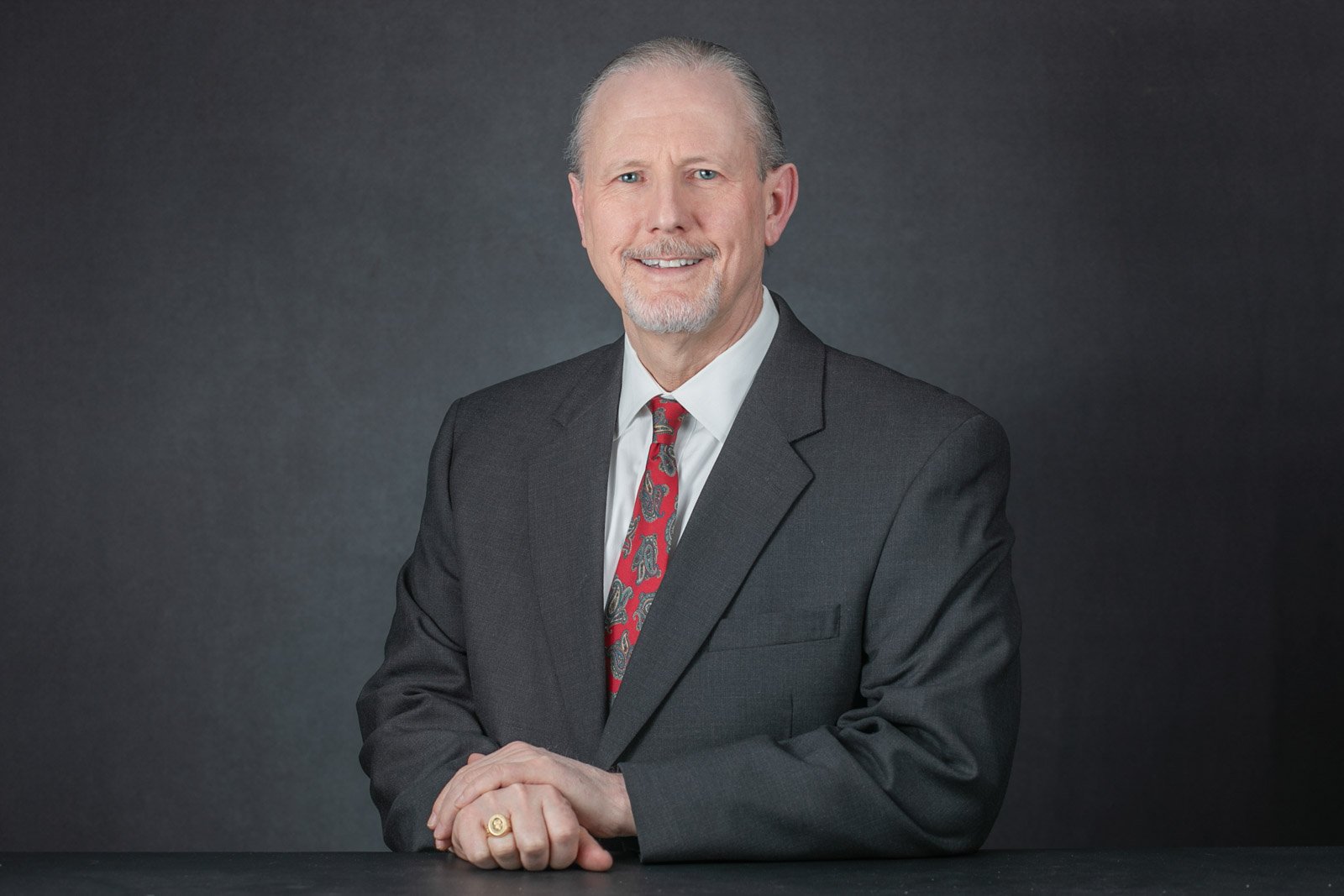 business-professional-headshot-executive-bloomfield-hills Professional headshot of a business executive sitting at a dest wearing a dark grey suit and a red tie. The background is a studio canvas