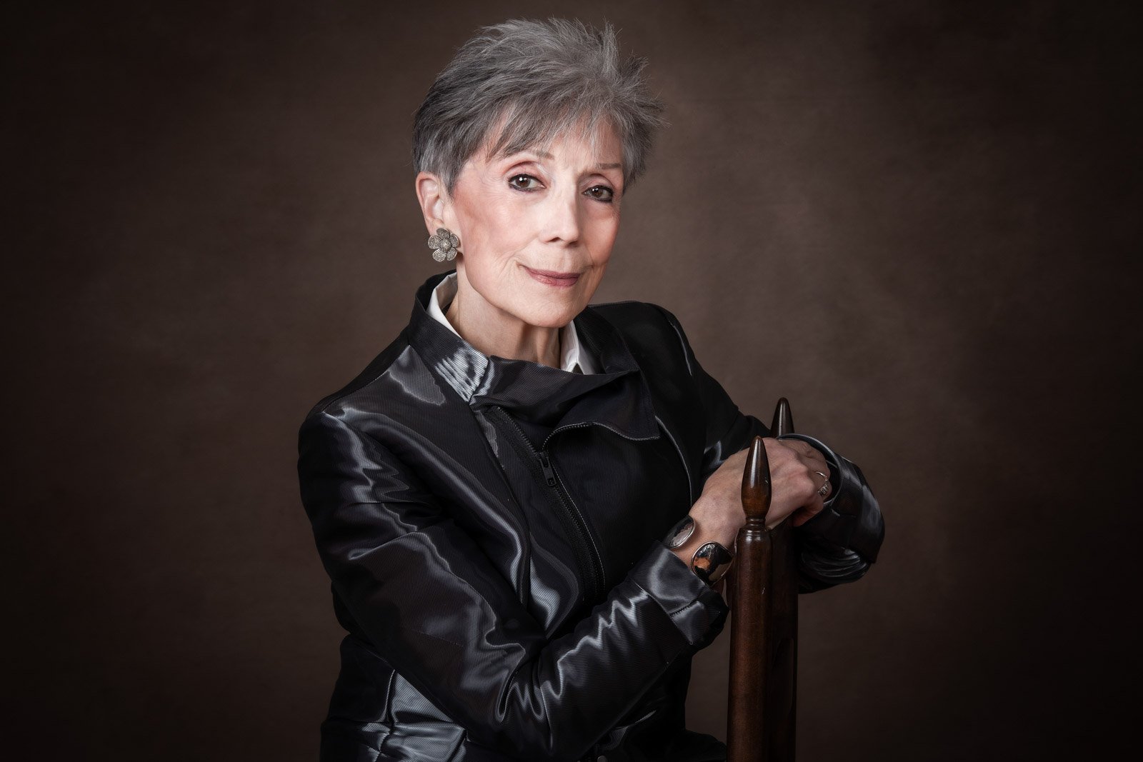 Mothers Day portrait of an elegant lady, sitting sideways on a dining chair. A studio portrait with brown tones, and she's wearing a leather jacket while sporting beautiful grey hair.