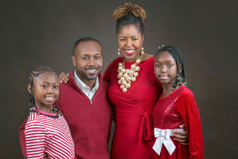 Mother poses with her husband and two young children girls, in a studio portrait session with a dark canvas background.