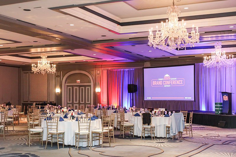 Corporate conference ballroom setup at the Ford’s Garage Brand Conference in Detroit with stage lighting, keynote presentation screen, and banquet seating