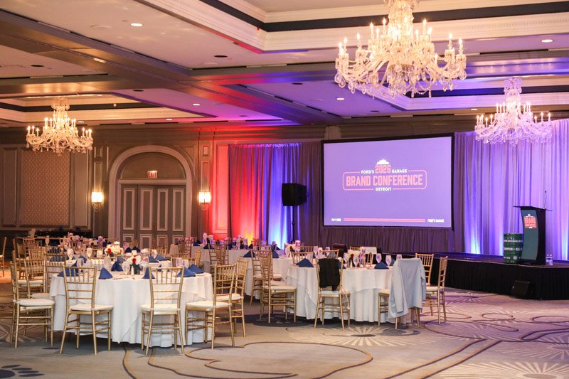 Ford's Garage corporate conference ballroom at The Henry Hotel in Dearborn, Michigan, featuring a branded stage, chandeliers, colorful lighting and banquet tables.
