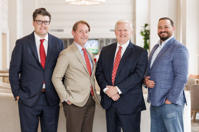 Four business executives pose for a professional group photograph in a bright hotel lobby.